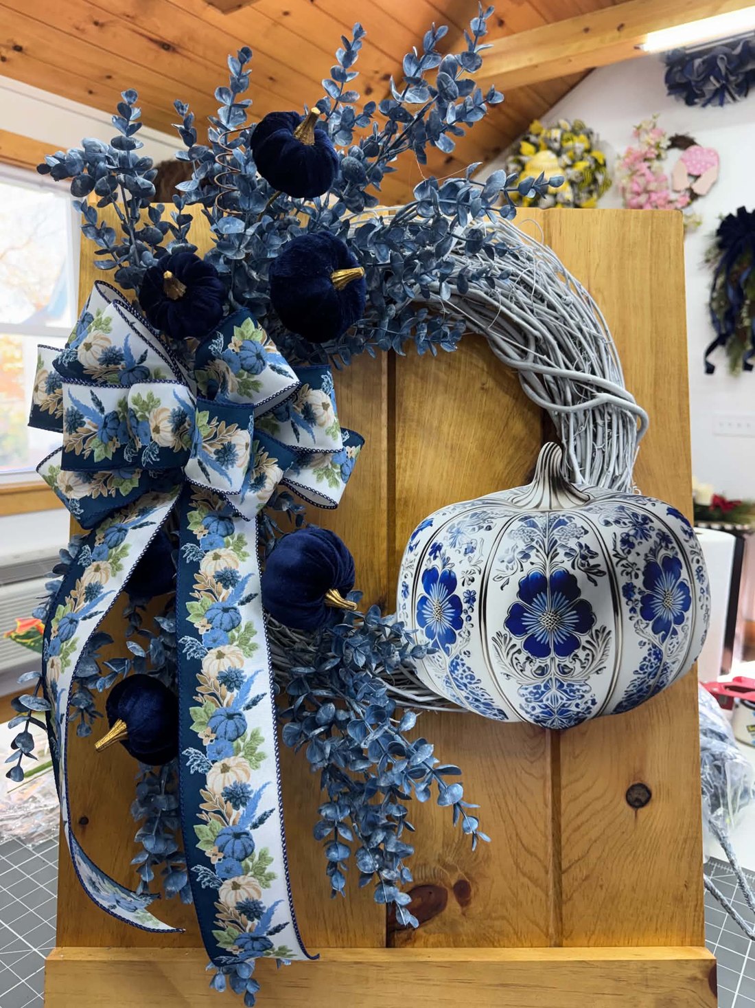 A whitewashed grapevine wreath with dusty-blue eucalyptus, navy velvet pumpkins, a blue-and-white floral bow with long tails, and a big blue-and-white porcelain-pattern pumpkin.