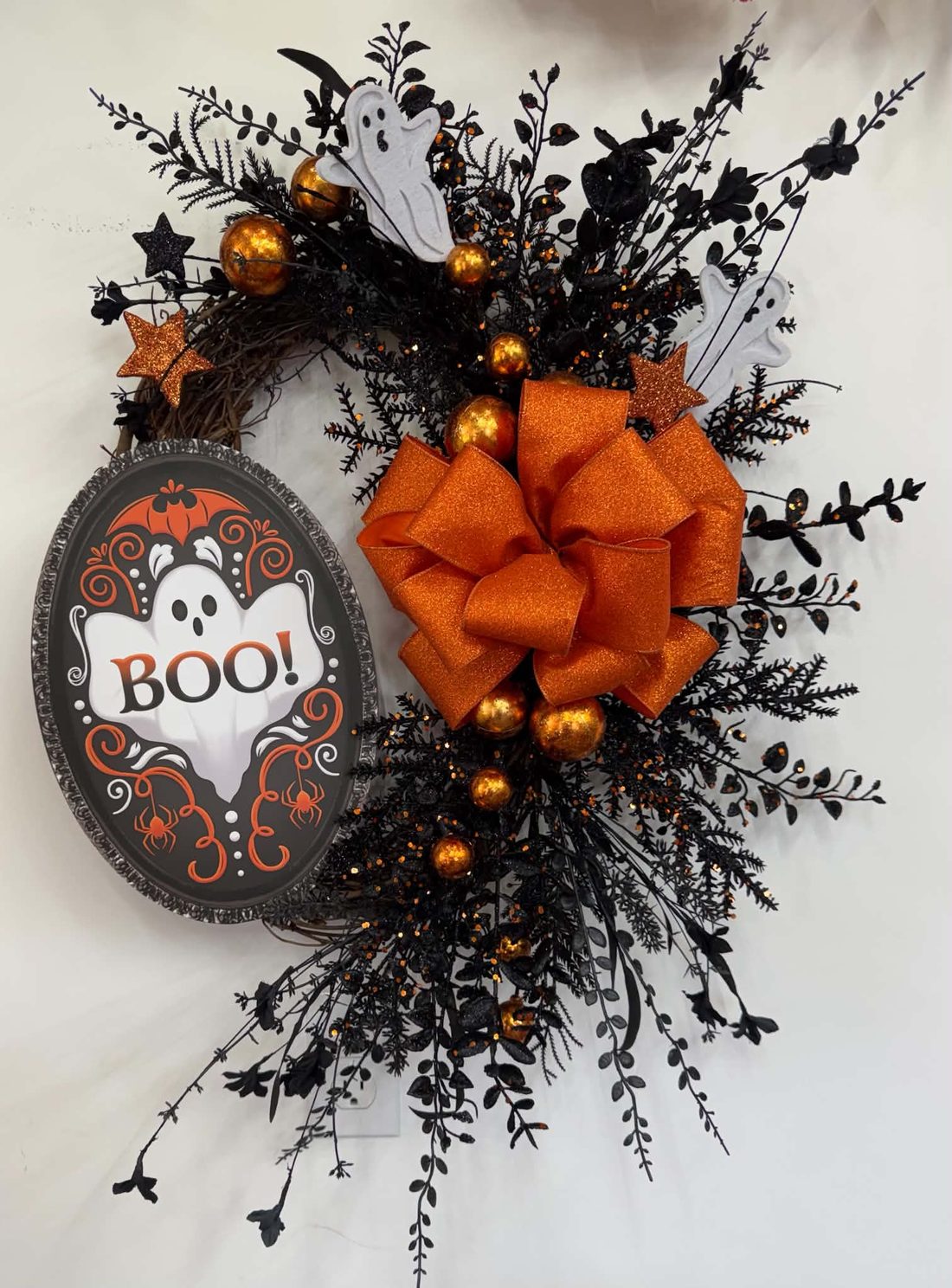 A framed 'BOO!' ghost sign on a crescent of black sprays with felt ghosts, copper balls, glitter stars, and a big orange glitter bow.
