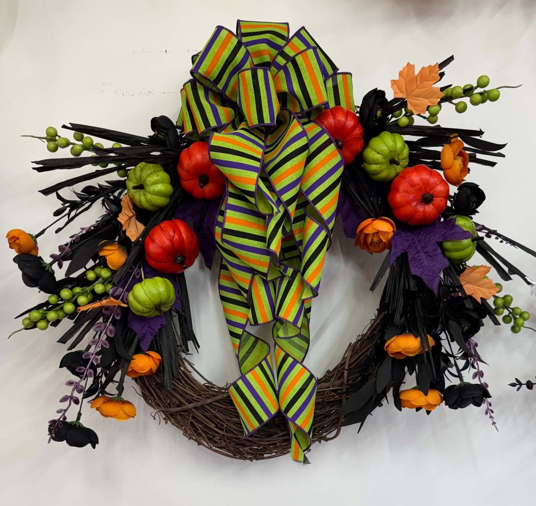 A grapevine wreath loaded with red and green pumpkins, orange ranunculus, and black sprays under a long striped bow in lime, purple, orange, and black.