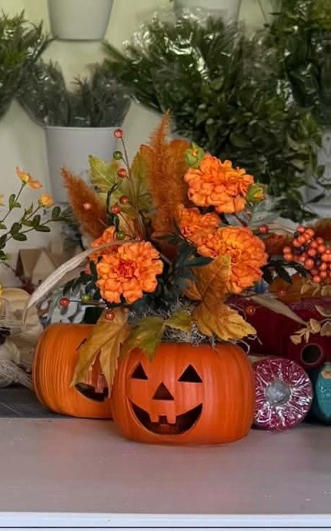 A smiling jack-o'-lantern planter overflowing with orange marigolds, autumn leaves, berries, and rust pampas plumes.