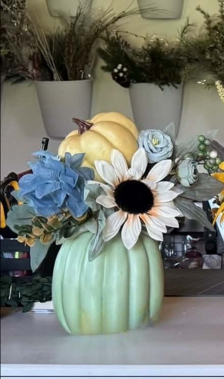 A sage-green pumpkin vase holding blue hydrangea, a cream sunflower, dusty-blue roses, and a little cream pumpkin on top.
